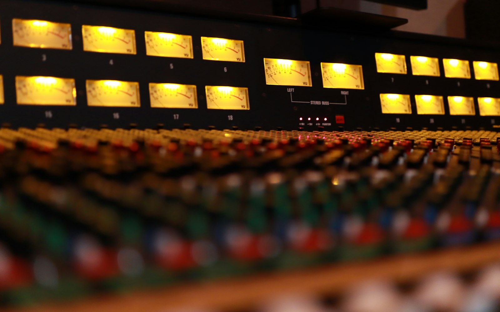 Close-up of a large analog mixing console with rows of glowing yellow VU meters above blurred faders and colored knobs in a dim studio