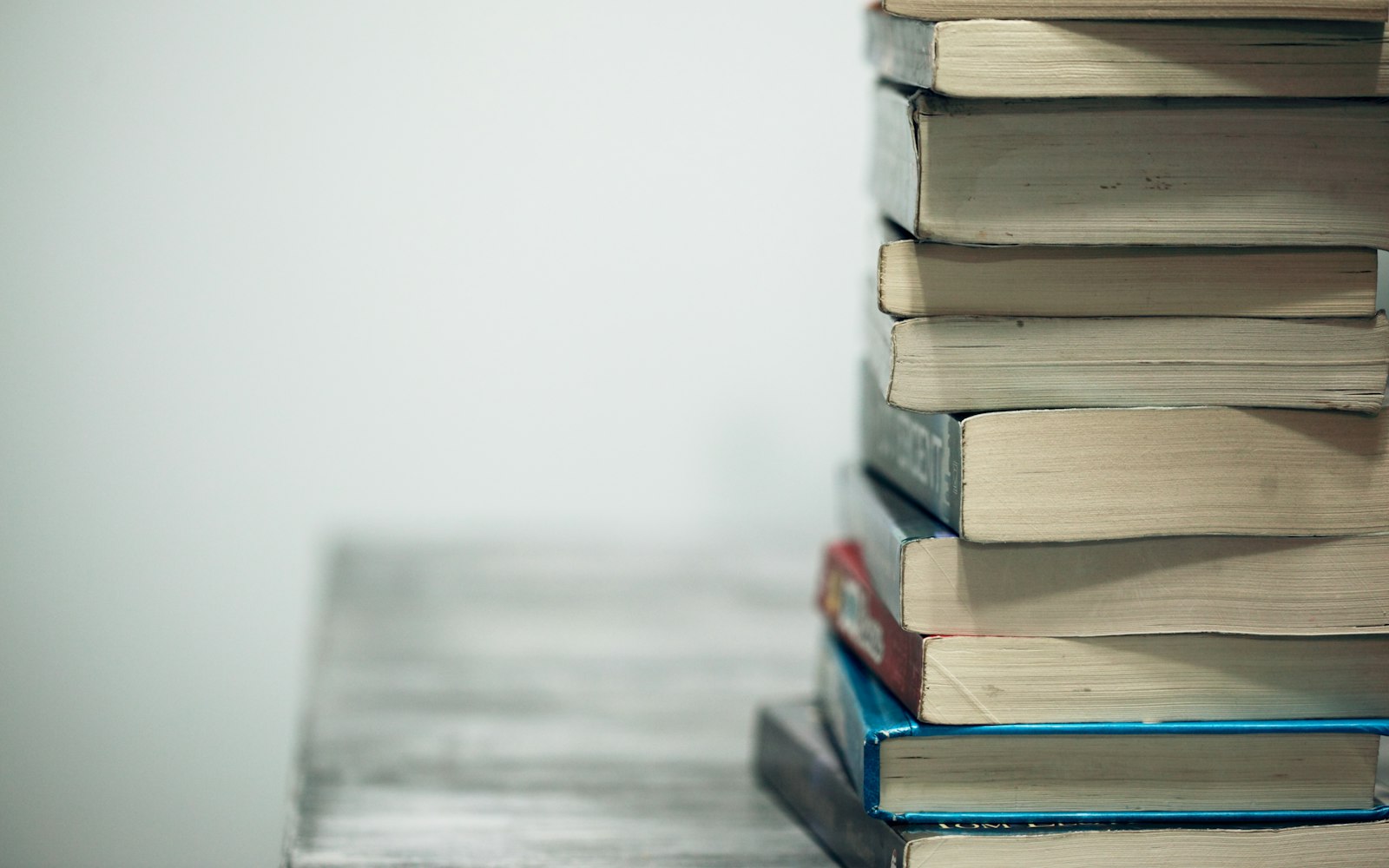 A tall stack of worn paperback books on a wooden table against a plain pale wall, with the page edges facing the camera