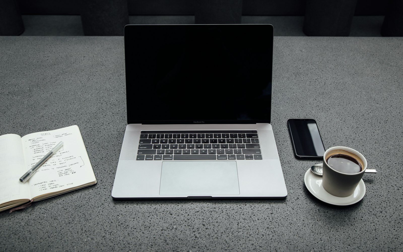 An open laptop on a grey desk between a notebook of handwritten notes with a pen, a smartphone and a cup of black coffee