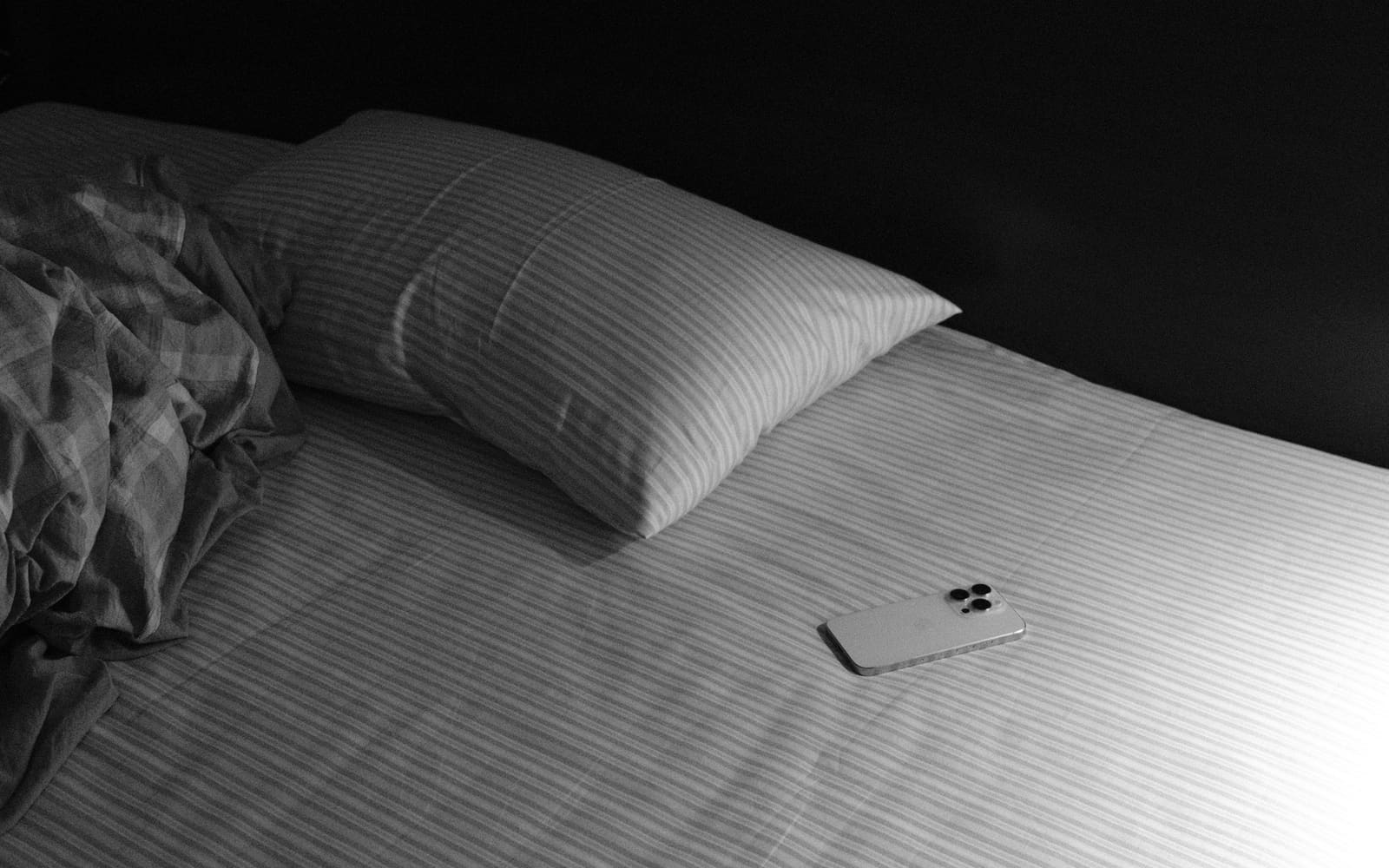A smartphone lies face down on an unmade bed with striped sheets and a rumpled checked duvet, in black and white