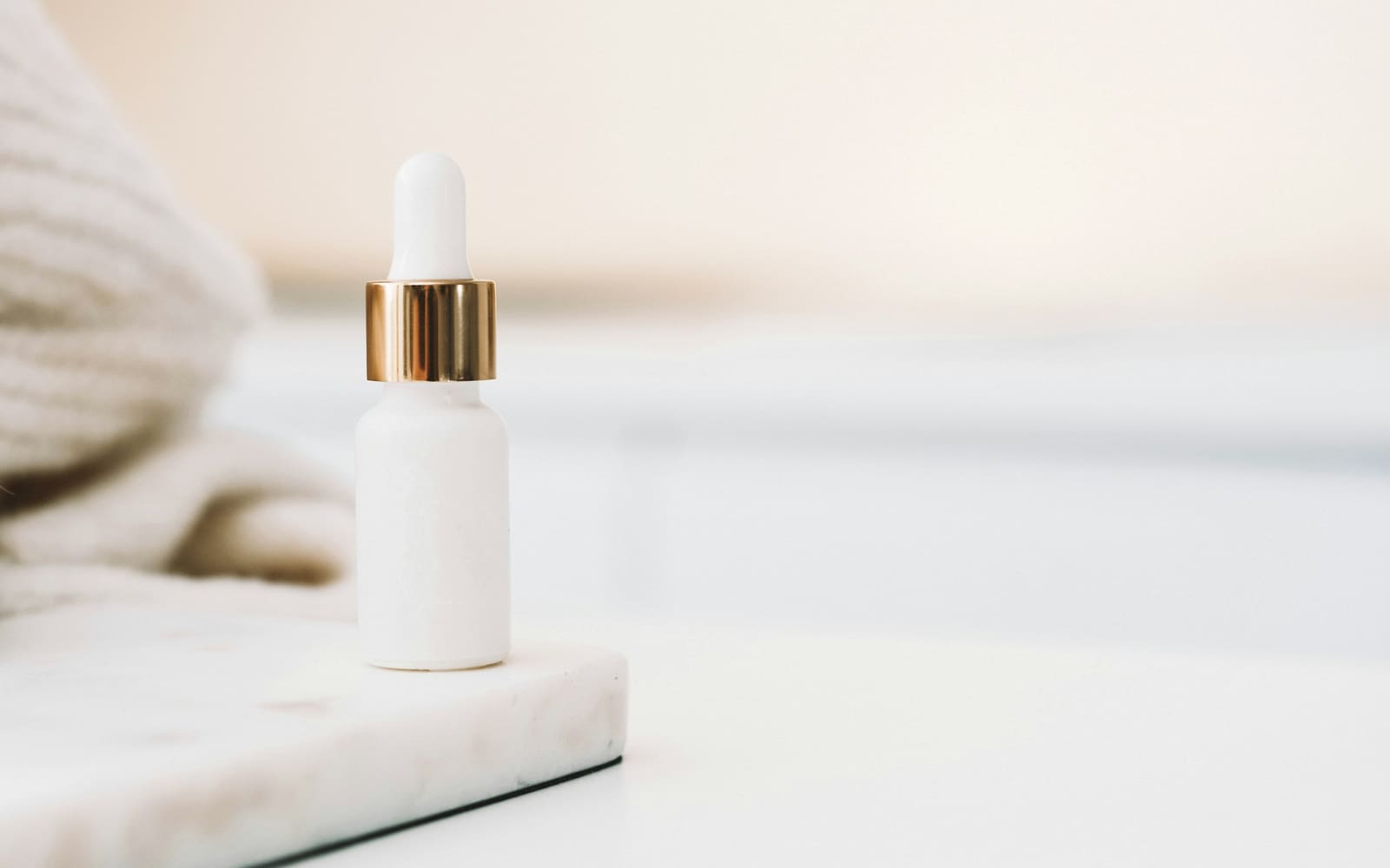A small white dropper bottle with a gold collar stands on a white marble tray beside a cream knit blanket in soft light