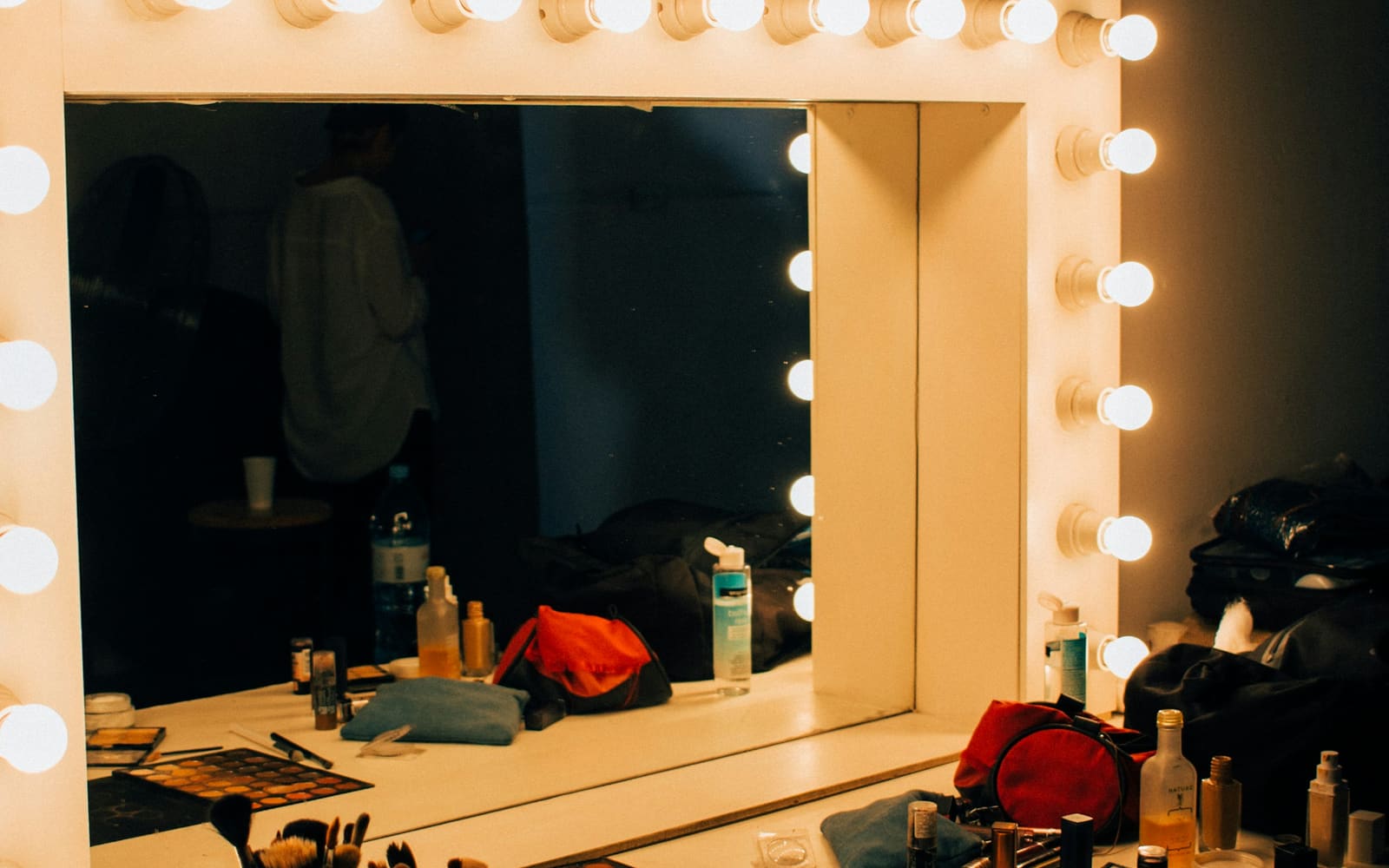 A makeup vanity mirror framed by glowing bulbs, with brushes, palettes and bottles on the counter and a blurred figure reflected in the glass
