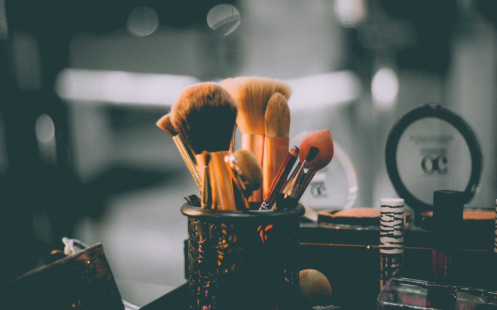 Makeup brushes with copper handles stand in a dark filigree holder beside a round compact and lipsticks, against a softly blurred gray background