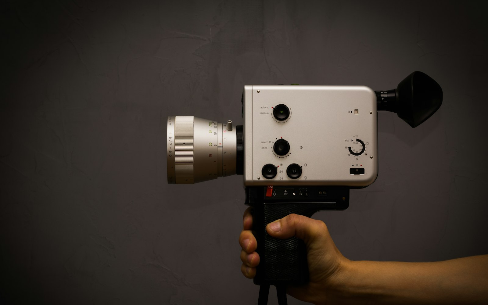 A hand gripping a vintage silver movie camera with a long zoom lens, dials and a black eyecup against a dark grey wall