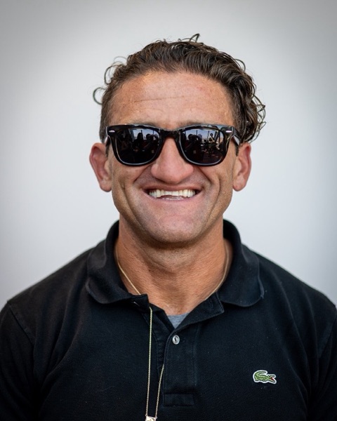 Casey Neistat smiling in black sunglasses and a black polo shirt against a light grey background