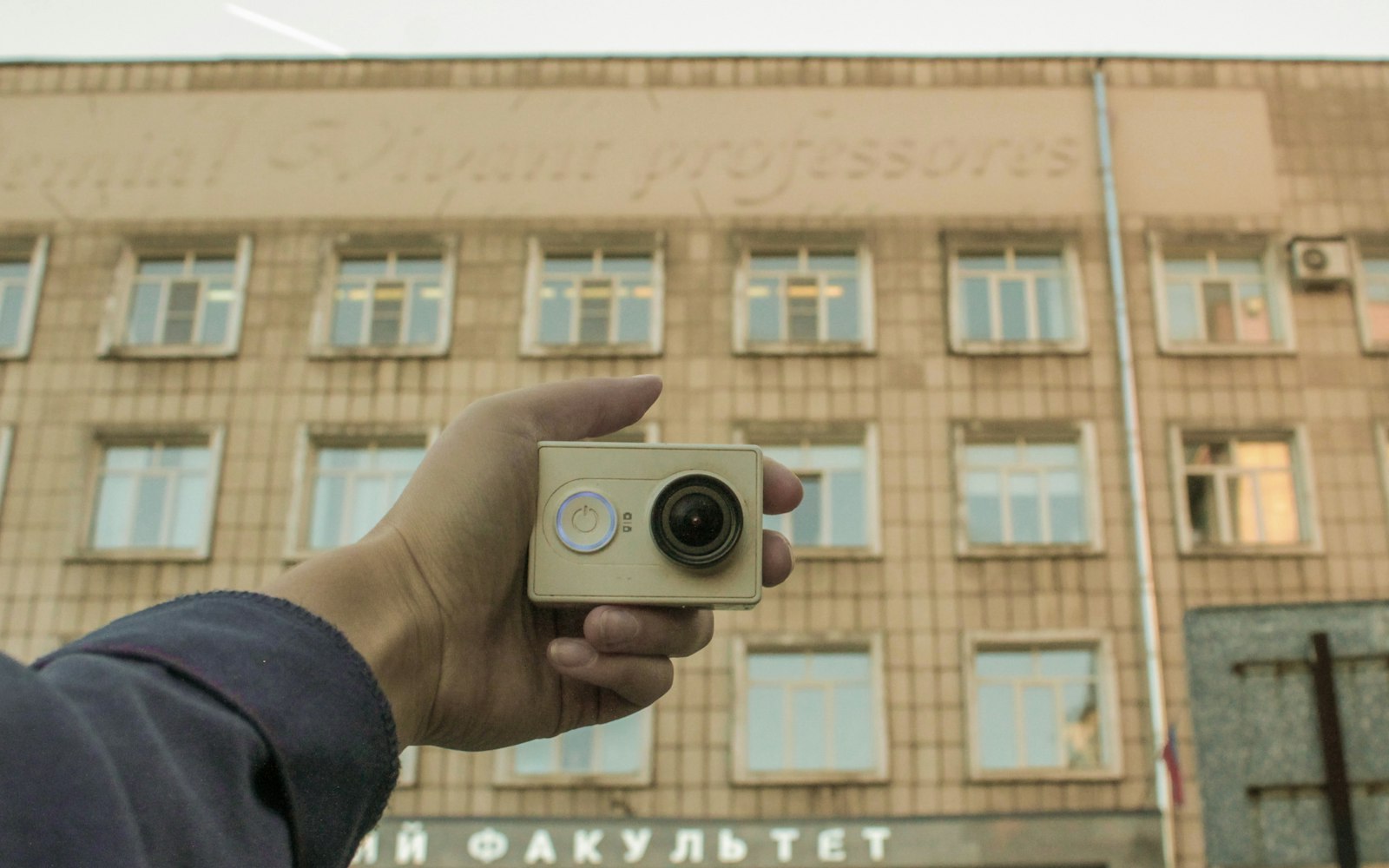 A hand holding a small action camera at arm’s length in front of a tiled multi-storey building with rows of windows