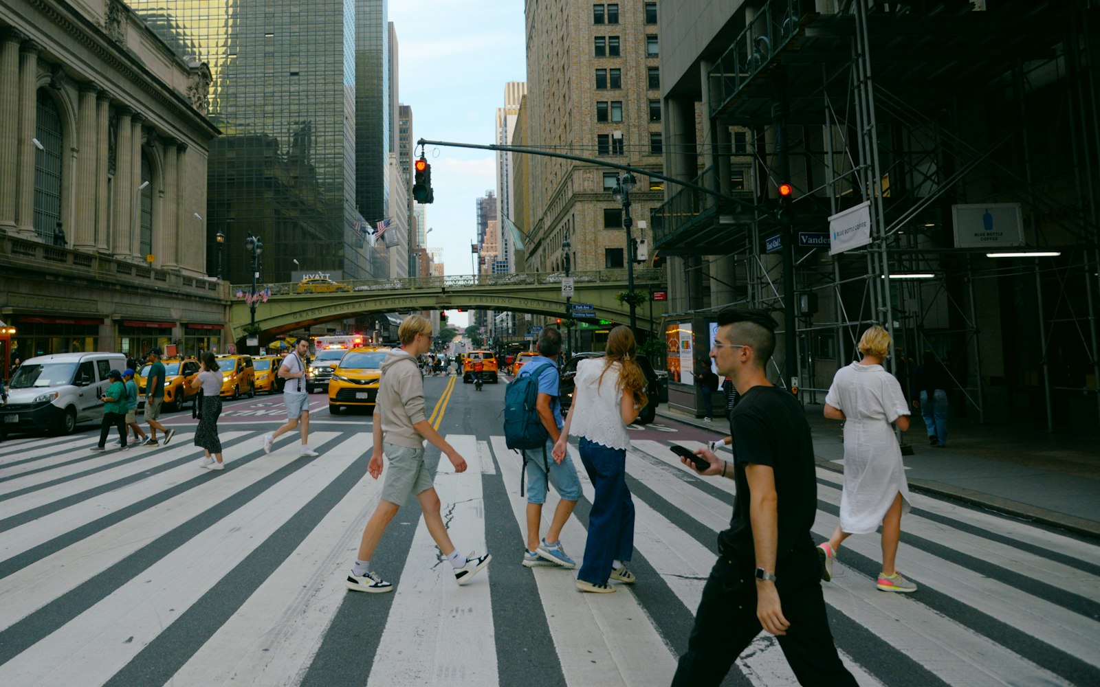 Pedestrians crossing a wide striped crosswalk on a busy New York street, with yellow taxis, skyscrapers and a stone viaduct behind them