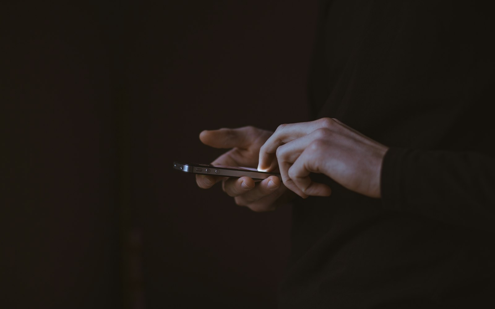 Two hands holding a smartphone in a dark room, one finger tapping the softly glowing screen