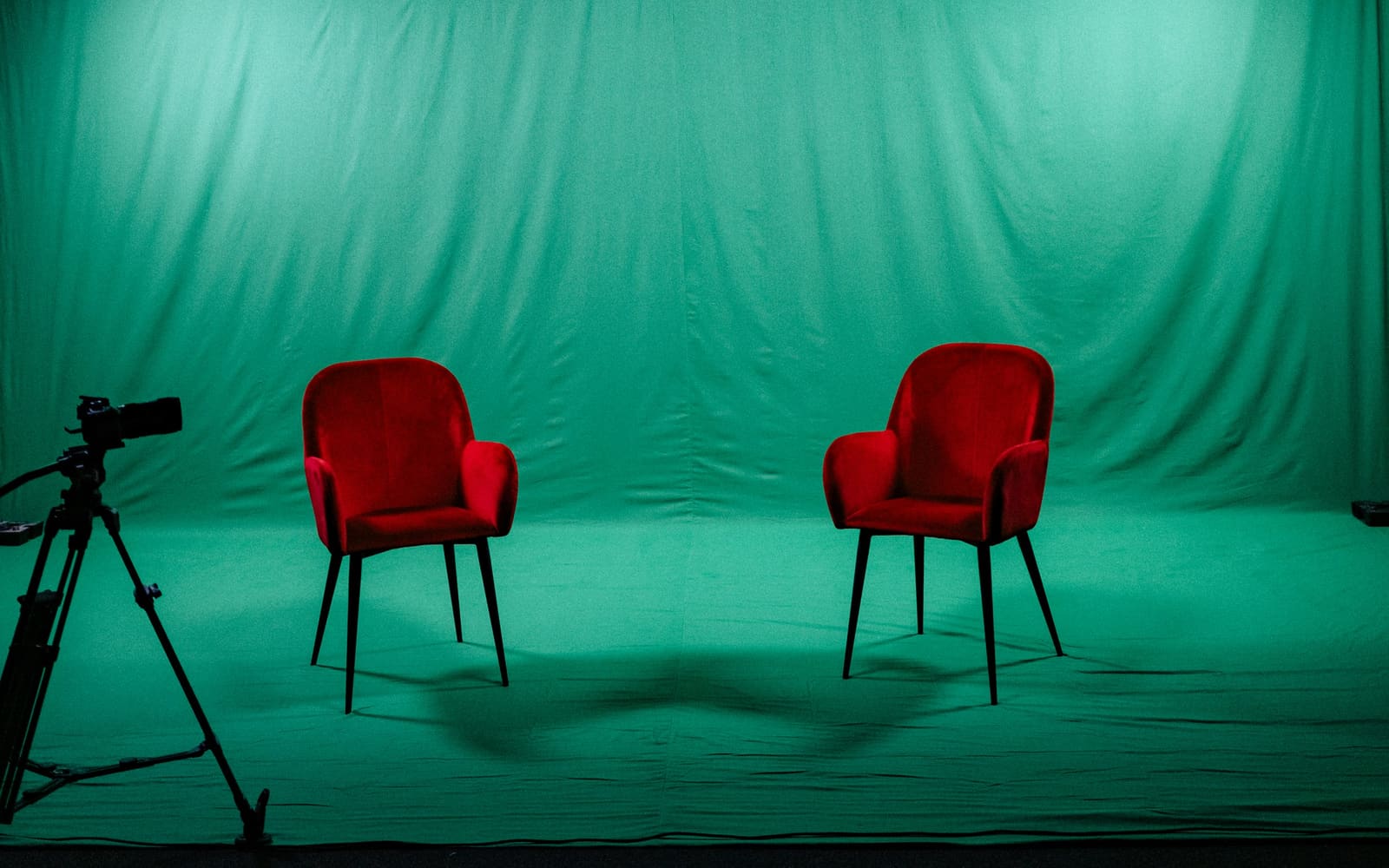 Two red velvet armchairs face each other on a green-screen studio floor, with a video camera on a tripod at the left edge