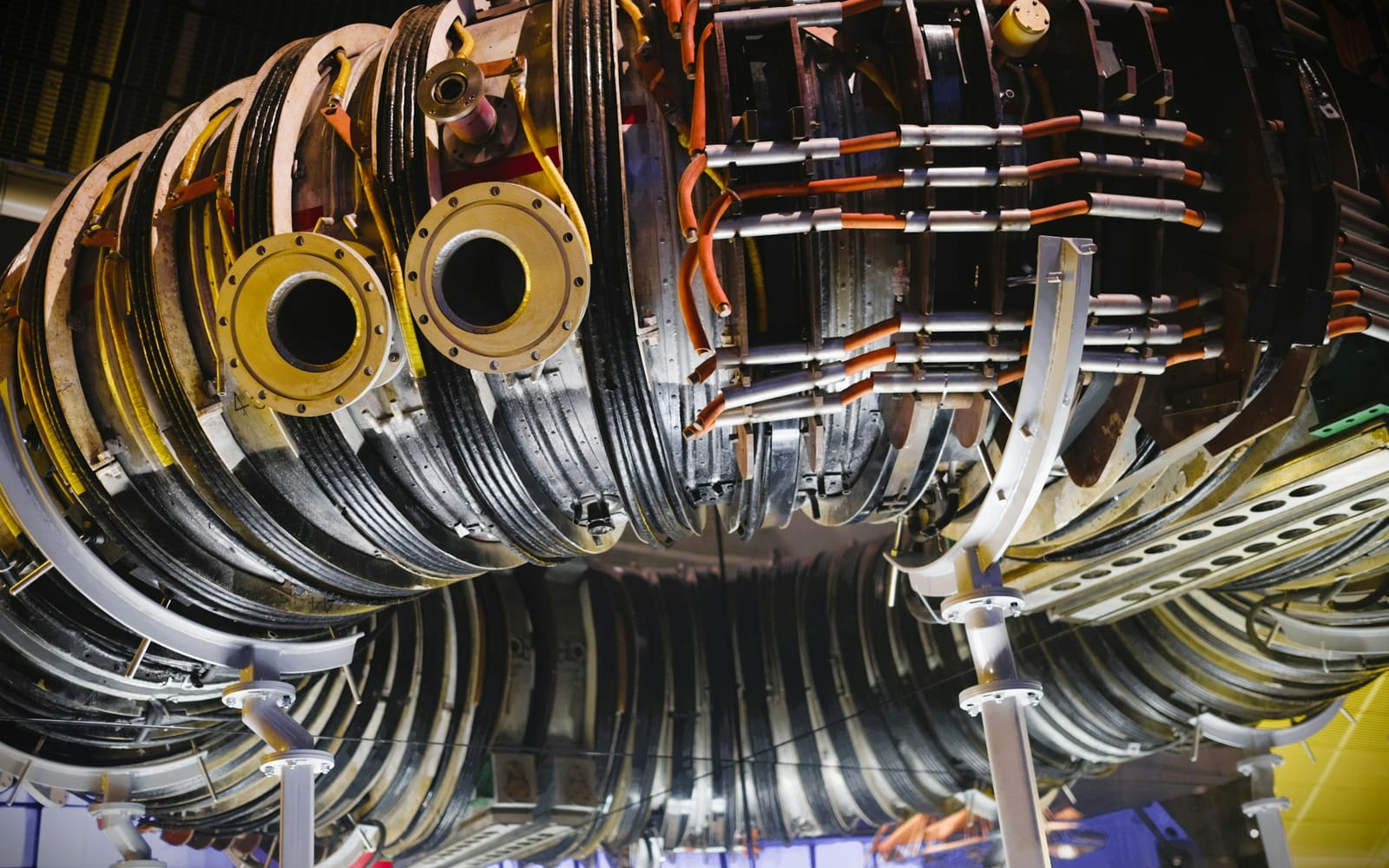 Rows of curved metal coil segments, brass flanges and orange-sheathed cables wrap a huge ring-shaped machine held up on white steel supports