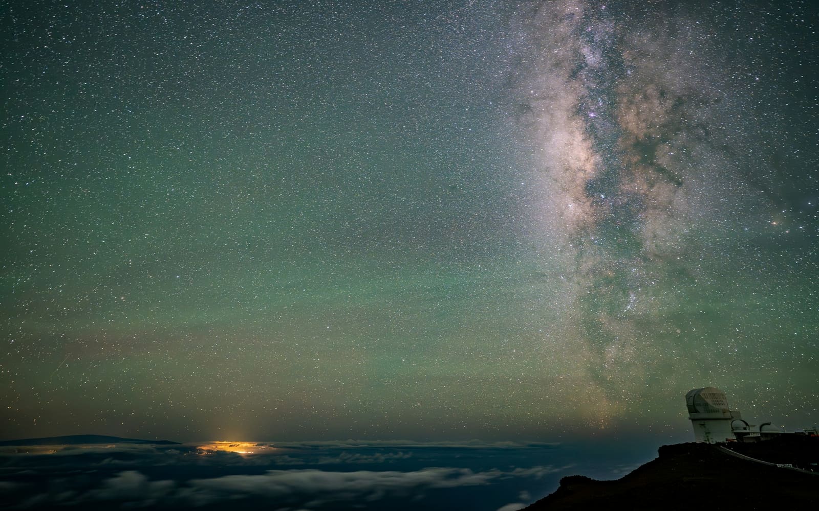 The Milky Way arcs over a white observatory dome on a dark mountain ridge, above a sea of clouds lit by a distant town