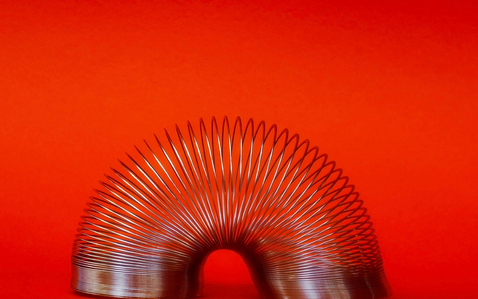 A metal spring toy bent into an arch, standing on both ends against a bright, plain red-orange background