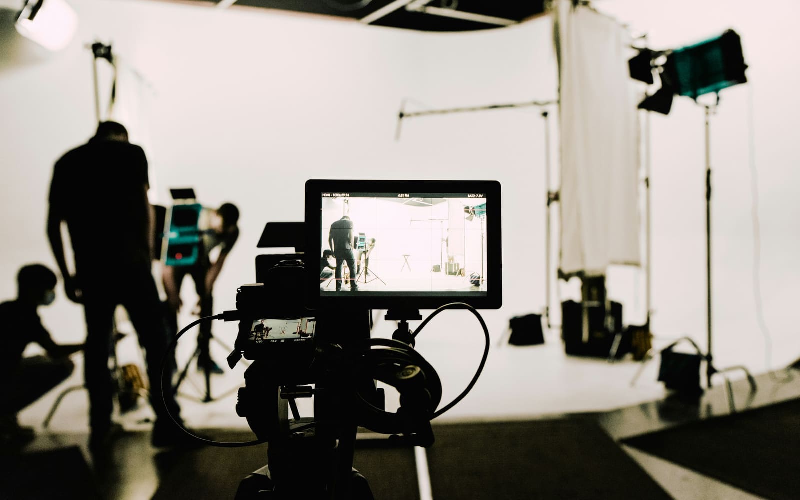 A camera monitor in the foreground shows a white studio set, while crew members in silhouette adjust lights and stands behind it