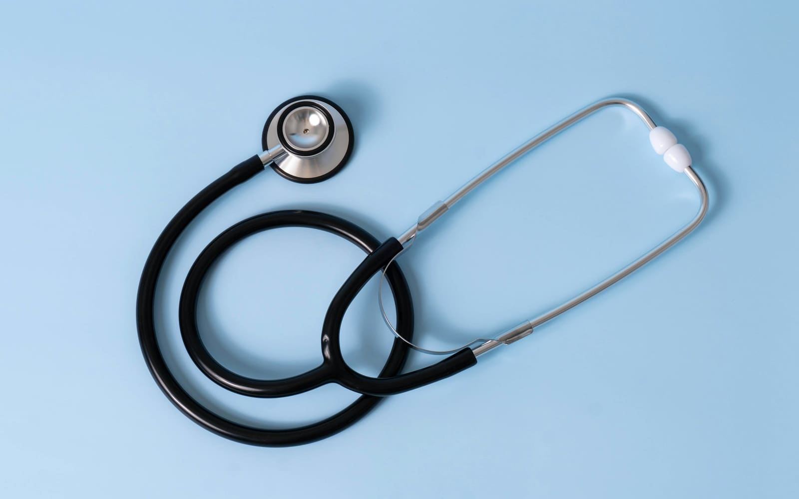 A black stethoscope with a silver chest piece and white ear tips lies coiled on a plain light blue background