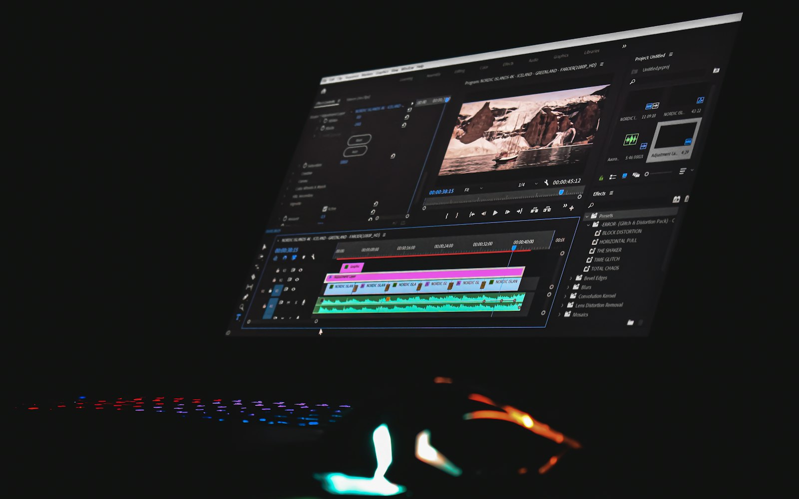 A monitor glowing in a dark room shows video editing software with a timeline, preview window and effects panel, above a backlit keyboard and mouse