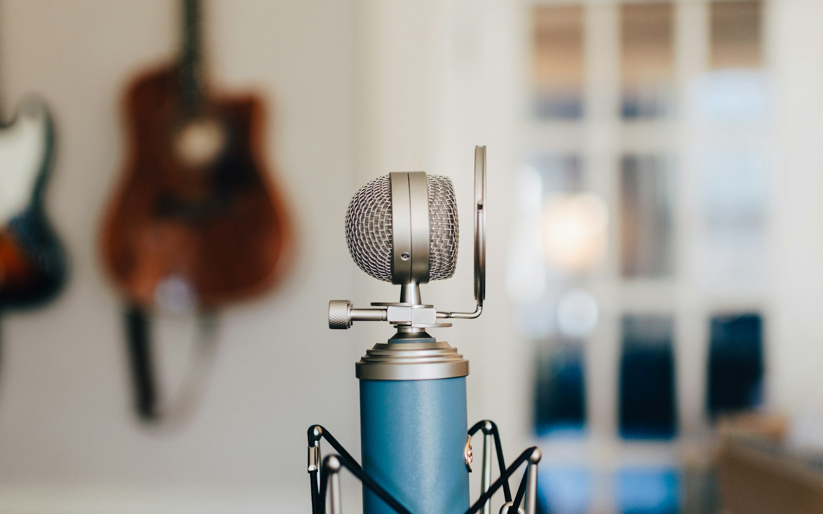 A blue studio microphone with a pop filter in a bright room, with two guitars hanging on the wall and a window blurred behind