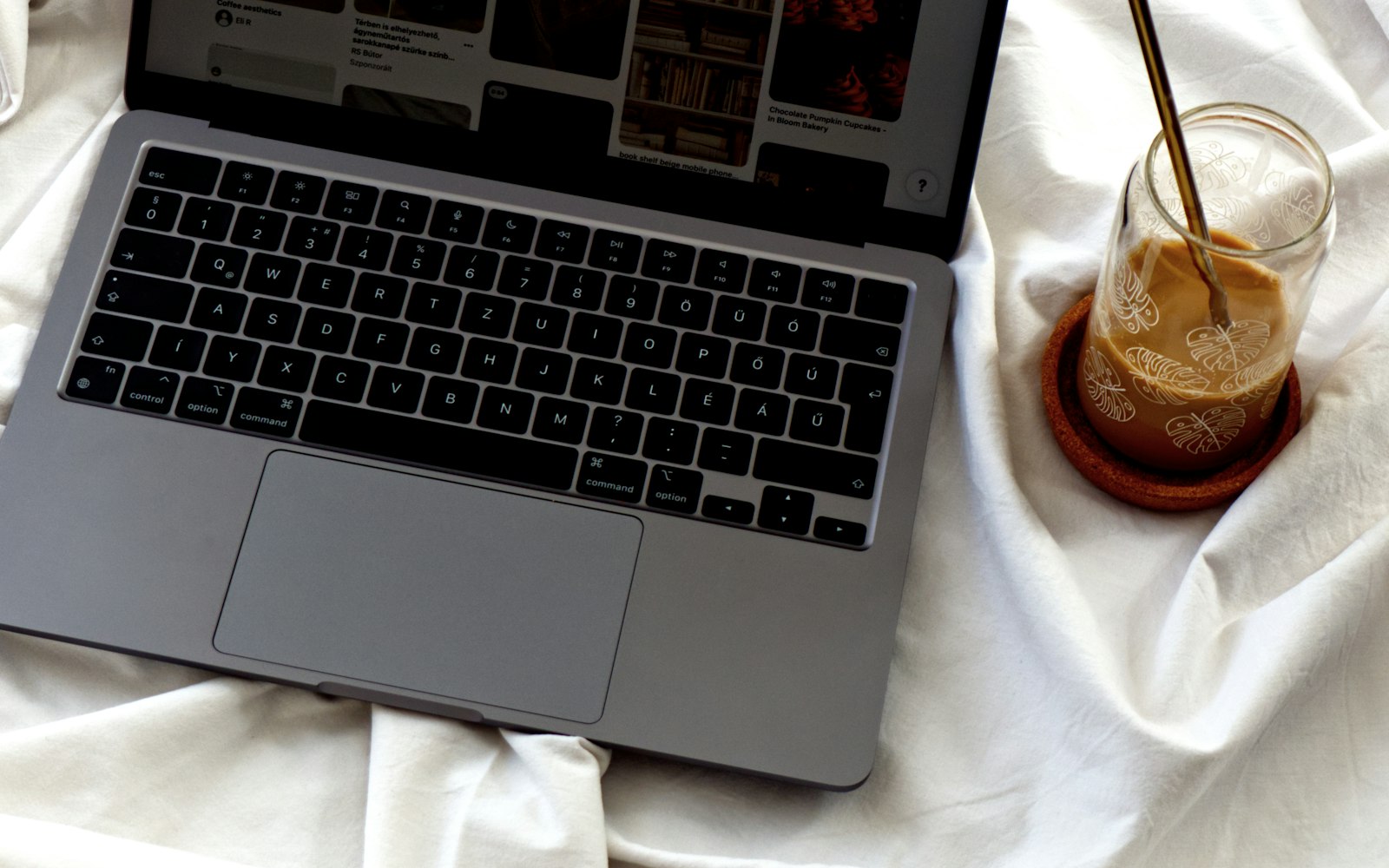 An open laptop on rumpled white bedsheets beside a glass of iced coffee with a gold straw, printed with leaf outlines and set on a cork coaster