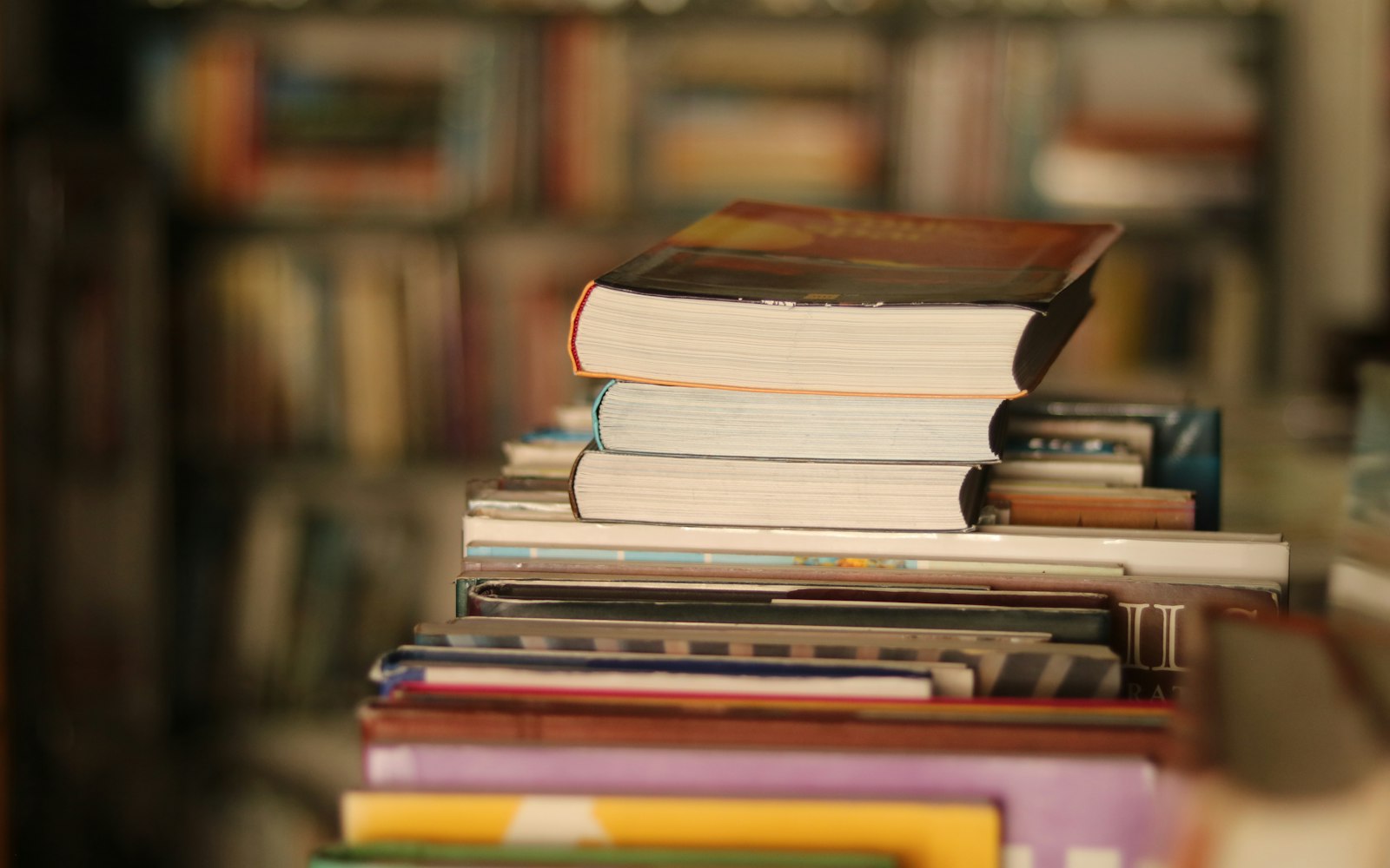A stack of thick paperback books on top of a row of hardcovers, with blurred bookshelves full of books in the background