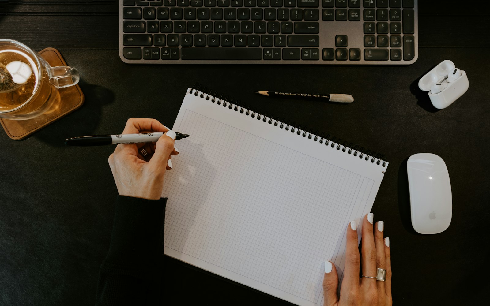 Overhead view of hands with a marker over a blank grid notebook on a dark desk, beside a keyboard, tea, earbuds and a mouse