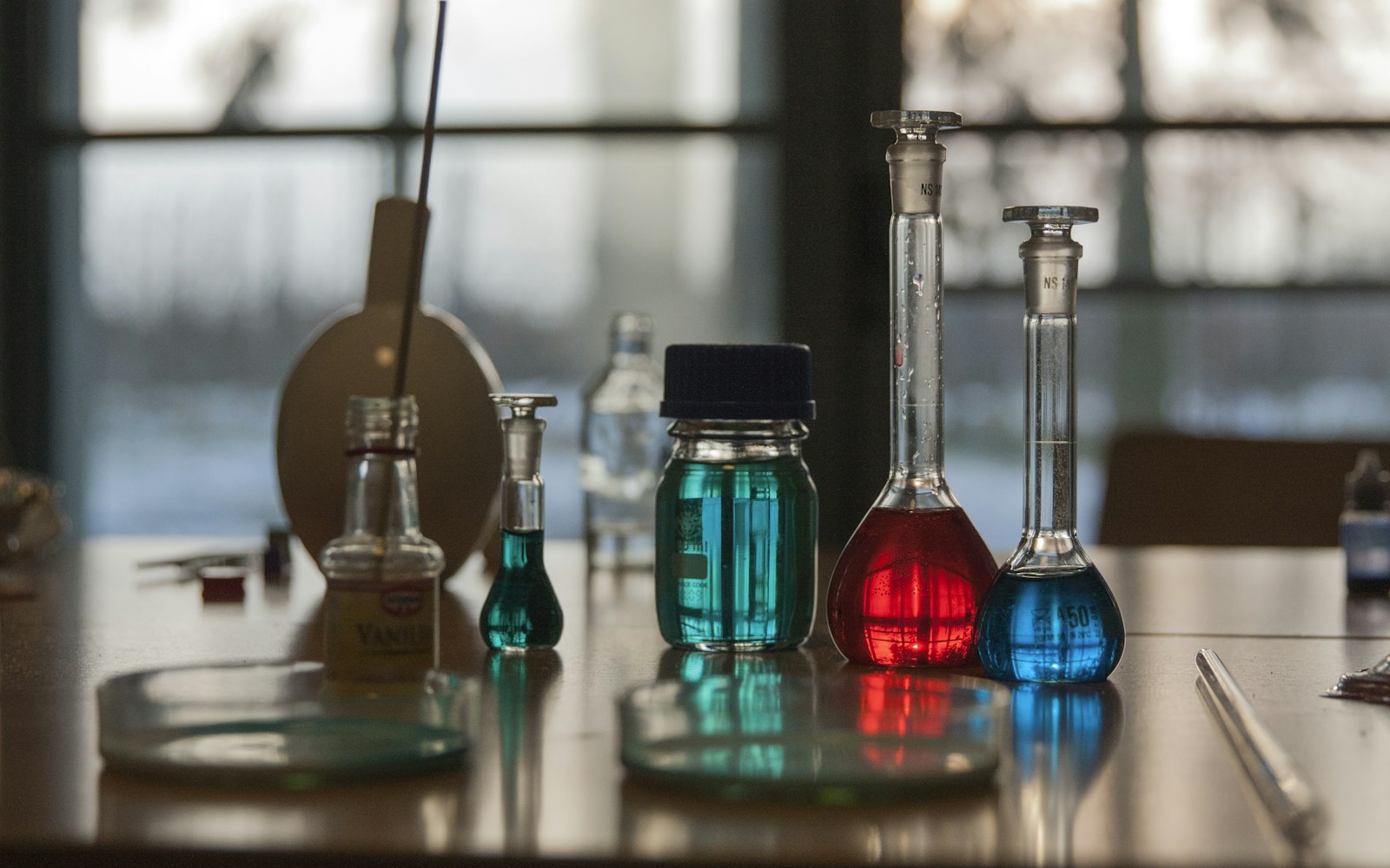 Glass flasks of red, blue and teal liquid, a lidded jar and petri dishes on a table in front of a large window