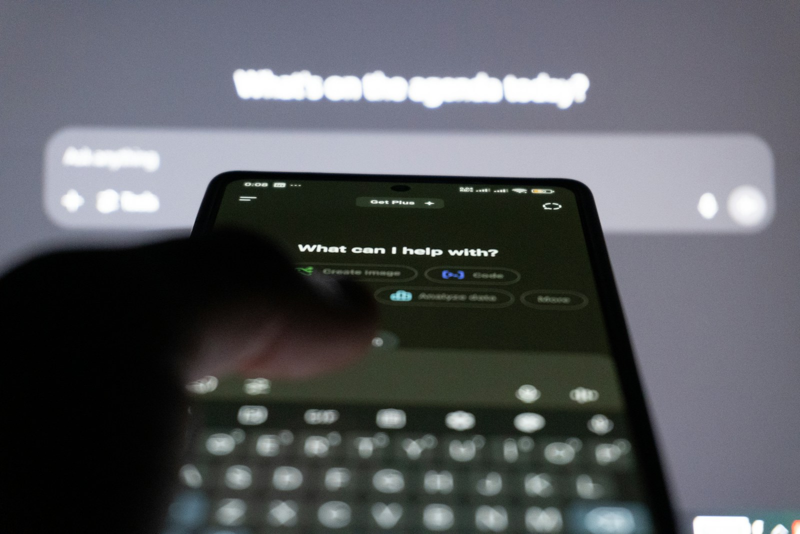 A finger taps a smartphone showing an AI chat app’s opening screen, with a large blurred screen behind showing the same assistant’s prompt box