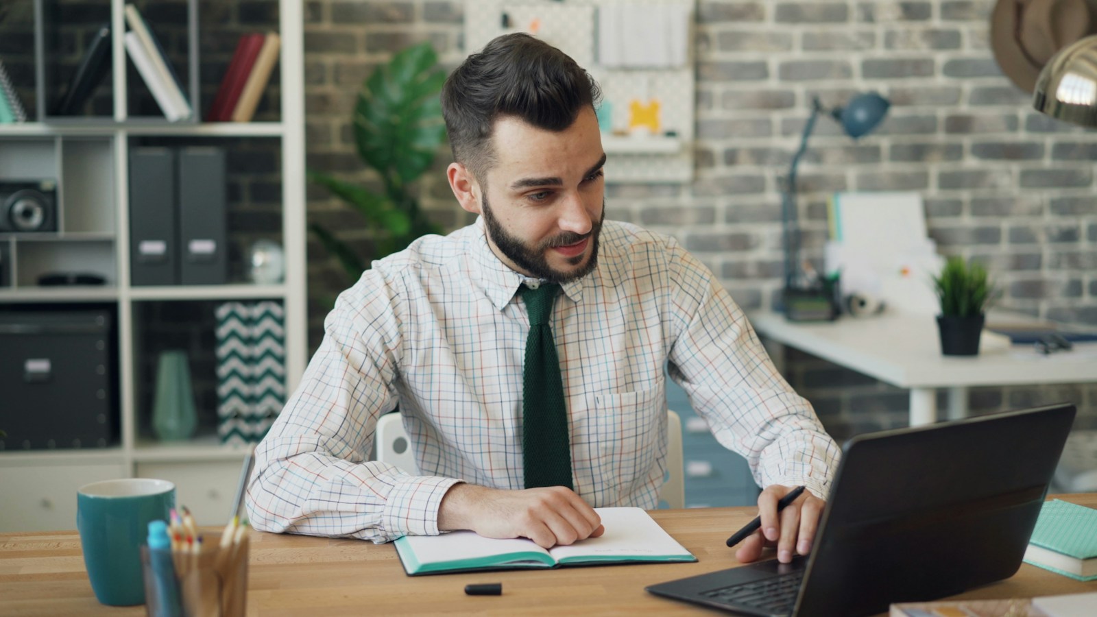 A bearded man in a checked shirt and green tie looks at his laptop with a doubtful half-smile, pen in hand over an open notebook