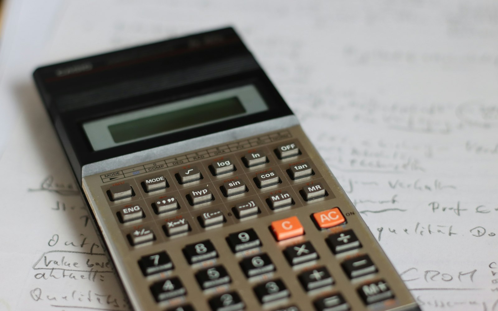 A worn scientific calculator with orange C and AC keys lies on a sheet of handwritten notes, the writing slightly out of focus around it