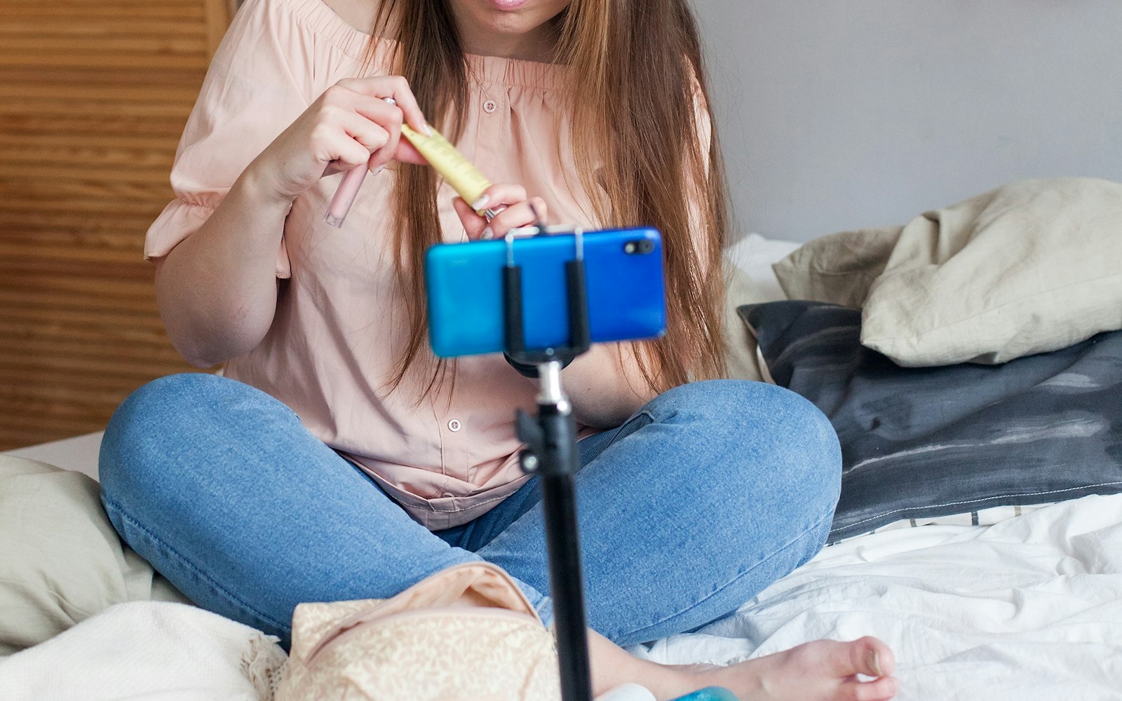 A woman sitting cross-legged on a bed holds a makeup product up to a blue smartphone clipped to a tripod, with her face cropped out of the frame