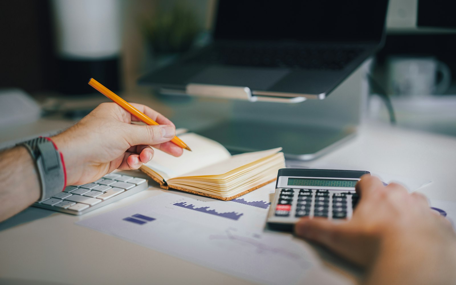 Close-up of one hand holding an orange pencil over an open notebook while the other uses a calculator, beside printed bar charts, a keyboard and a laptop on a stand