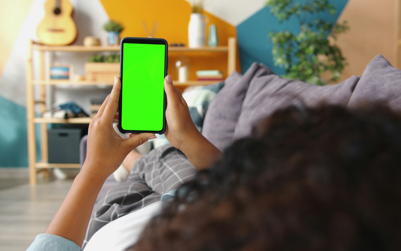 Over-the-shoulder view of a person lying on a grey sofa holding a smartphone upright with a blank green screen, in a living room with wooden shelves, plants and a guitar