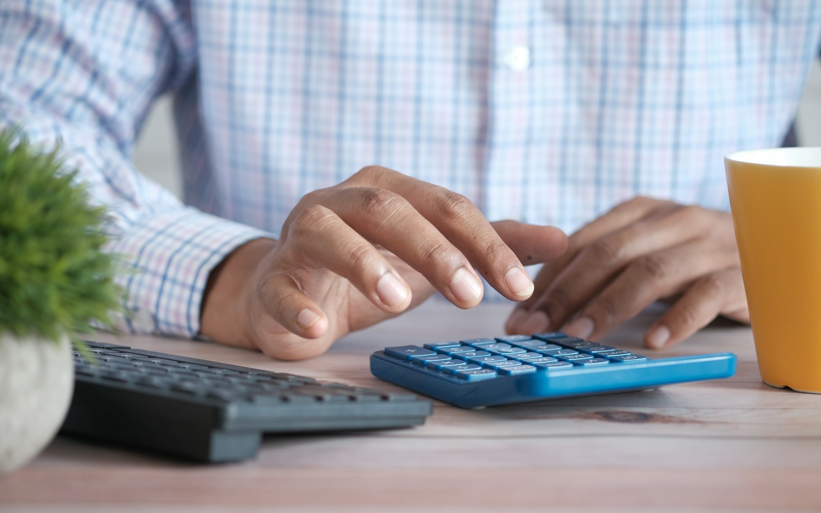 A person in a checked shirt typing on a blue calculator at a wooden desk, next to a keyboard, a small potted plant and a yellow mug