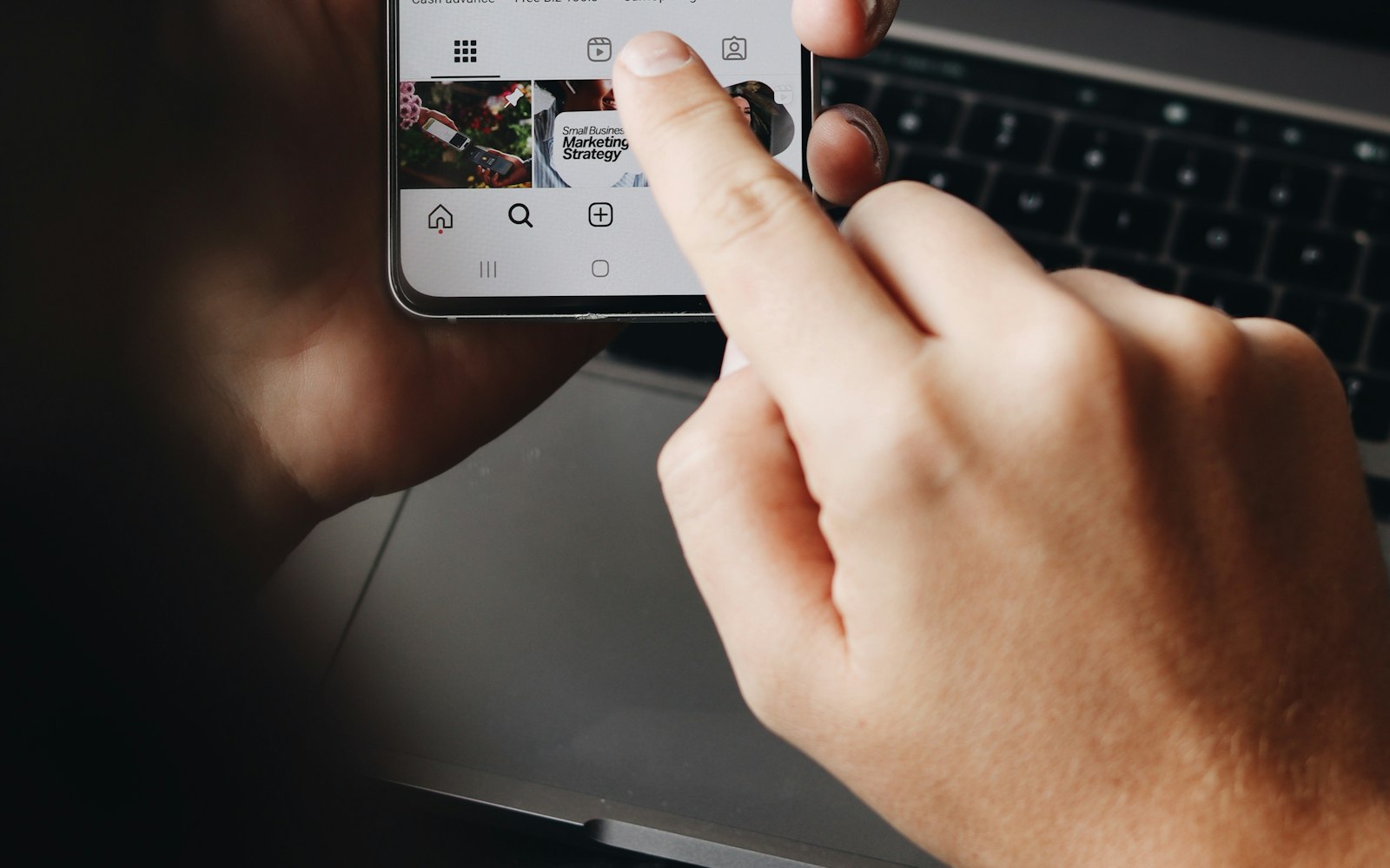 Close-up of a hand tapping a smartphone showing a social media profile grid with a small business marketing post, held above a laptop keyboard