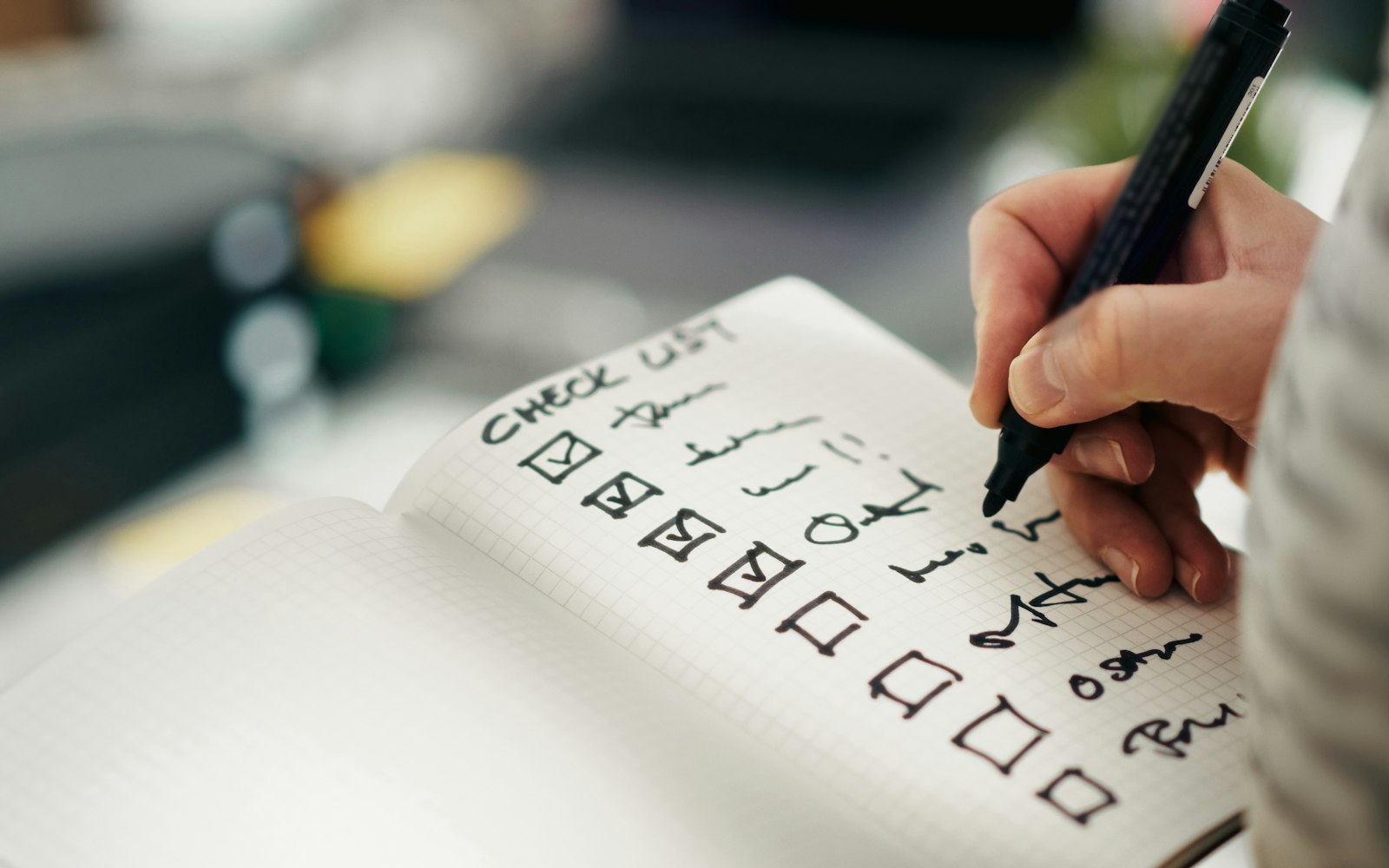 A hand writing with a black marker in a grid-paper notebook, ticking boxes on a handwritten checklist headed “check list”, with a blurred desk behind