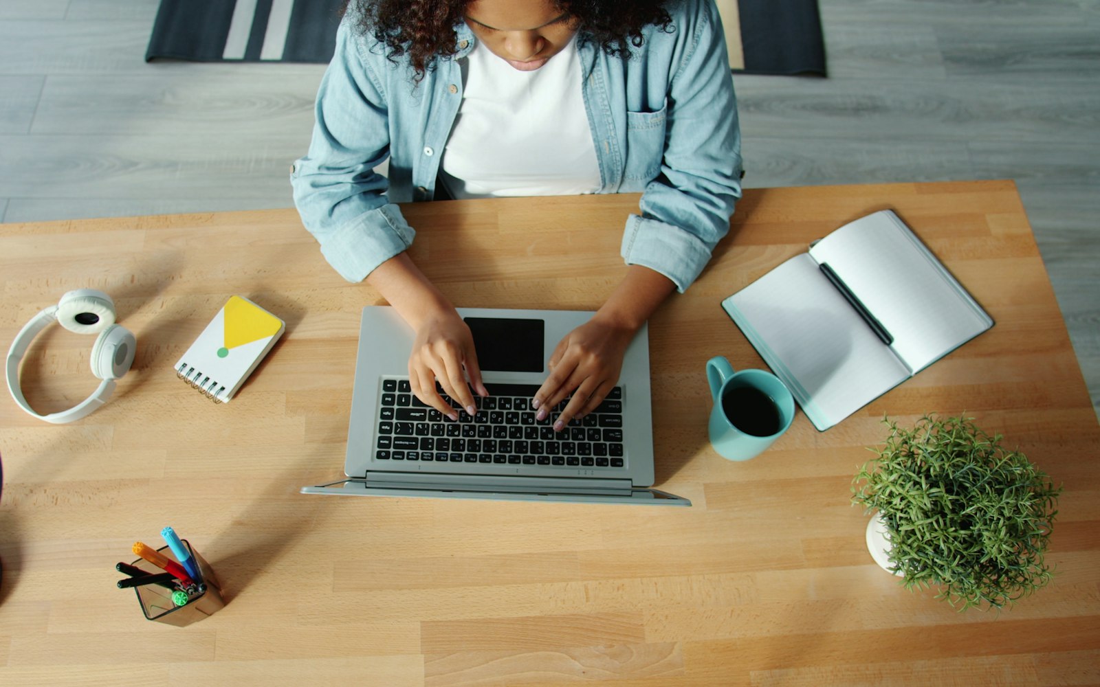 Overhead view of a woman in a denim shirt typing on a laptop at a wooden desk, with headphones, a notebook, a mug of coffee and a potted plant around it