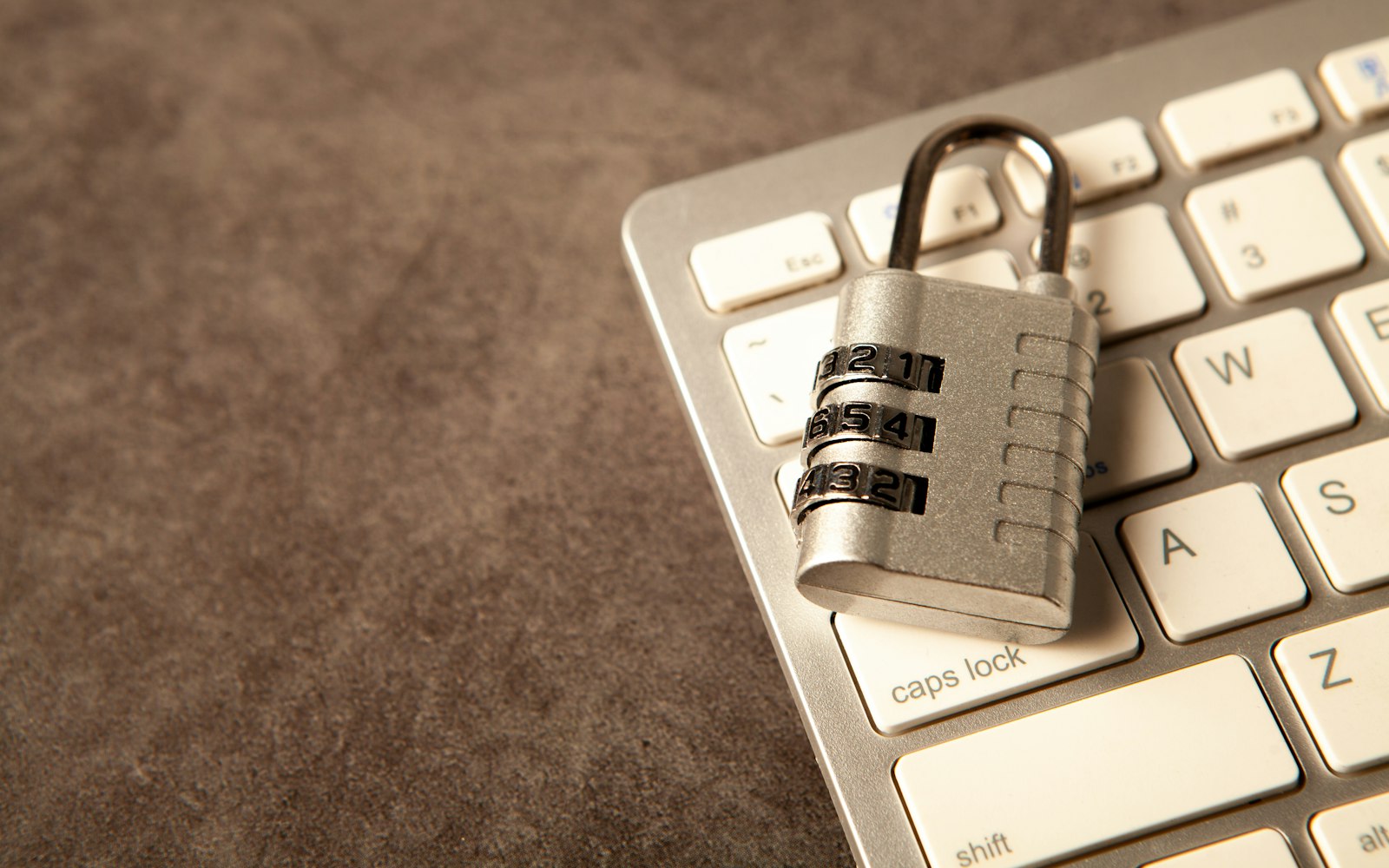 A silver combination padlock resting on the corner of a white computer keyboard, beside the caps lock and Escape keys, on a brown surface