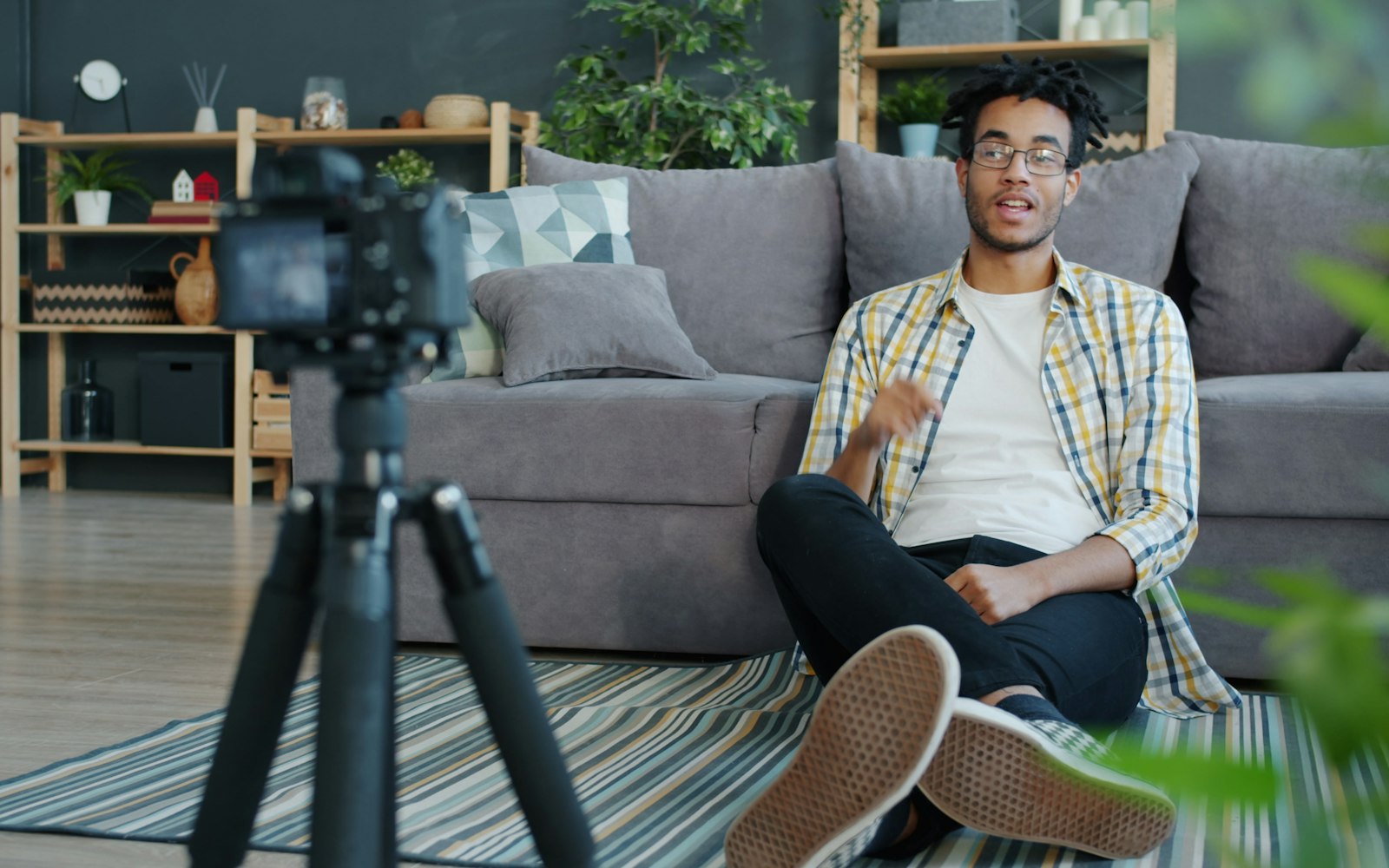 A young man in glasses and a plaid shirt sits on a striped rug in front of a grey sofa, talking to a camera on a tripod in his living room
