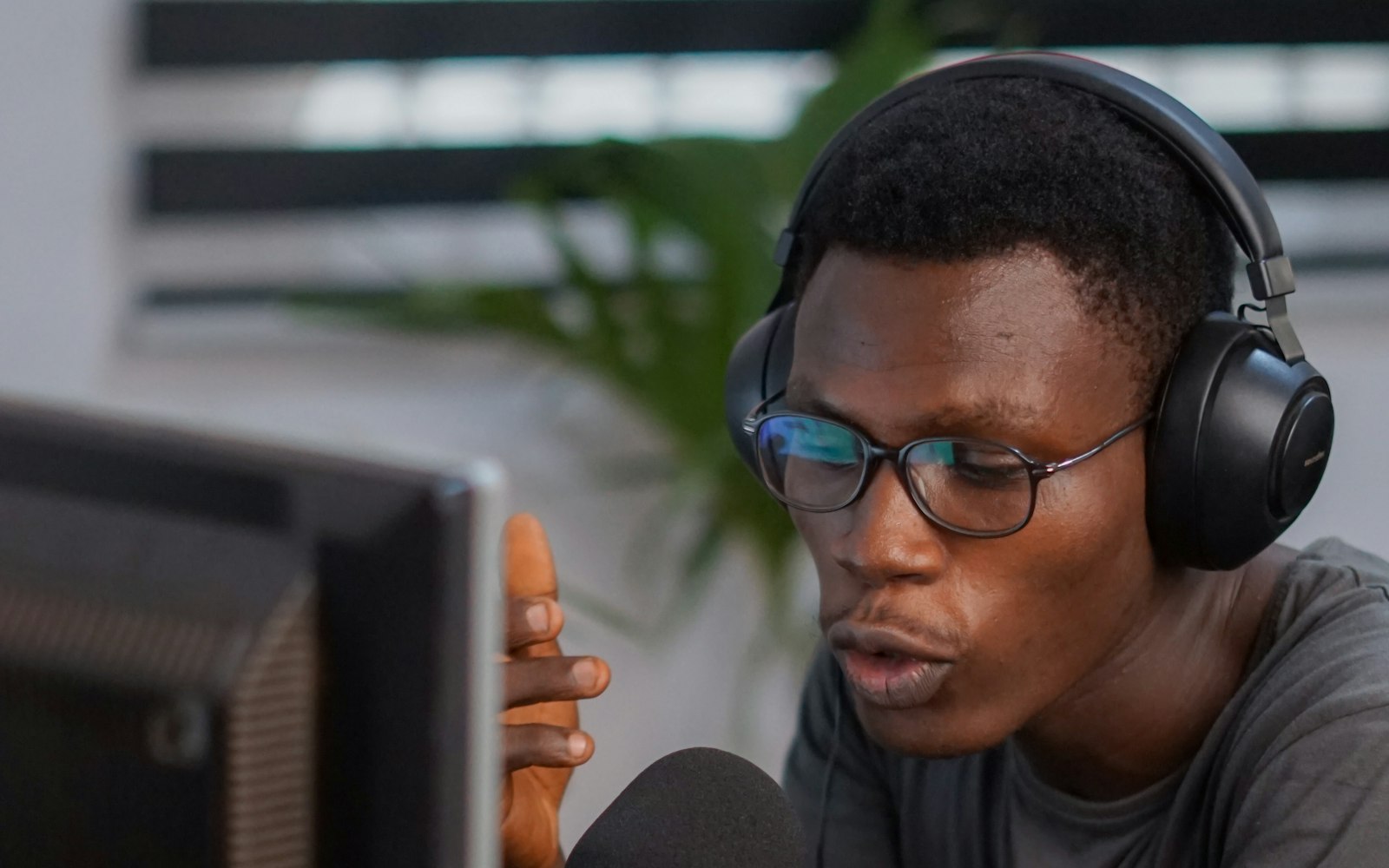A man in glasses and over-ear headphones speaks into a microphone while looking at a computer monitor, gesturing with one hand, with a plant behind him