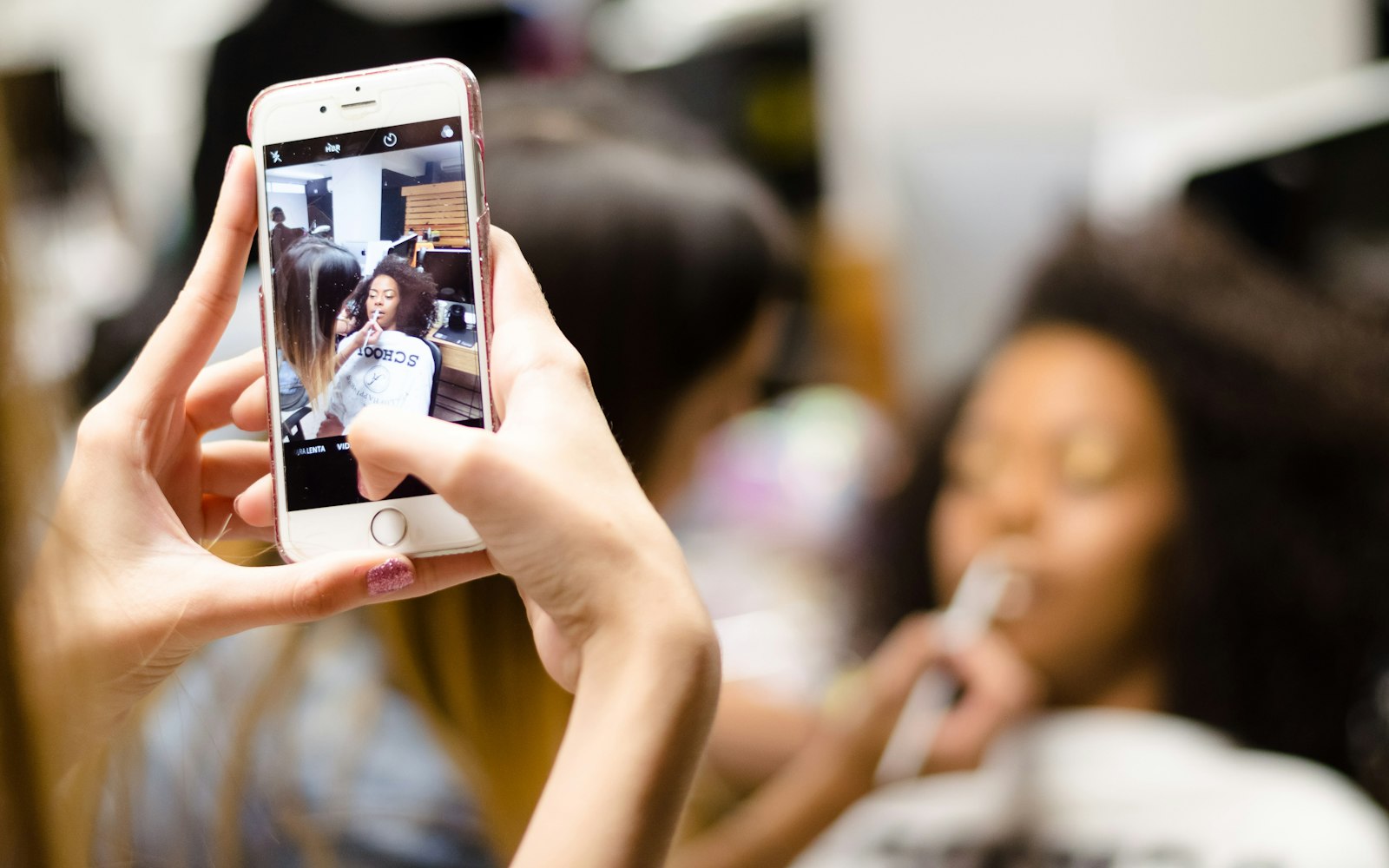 A hand holding a smartphone filming a makeup artist applying lip gloss to a woman in a salon chair, shown on screen and blurred behind