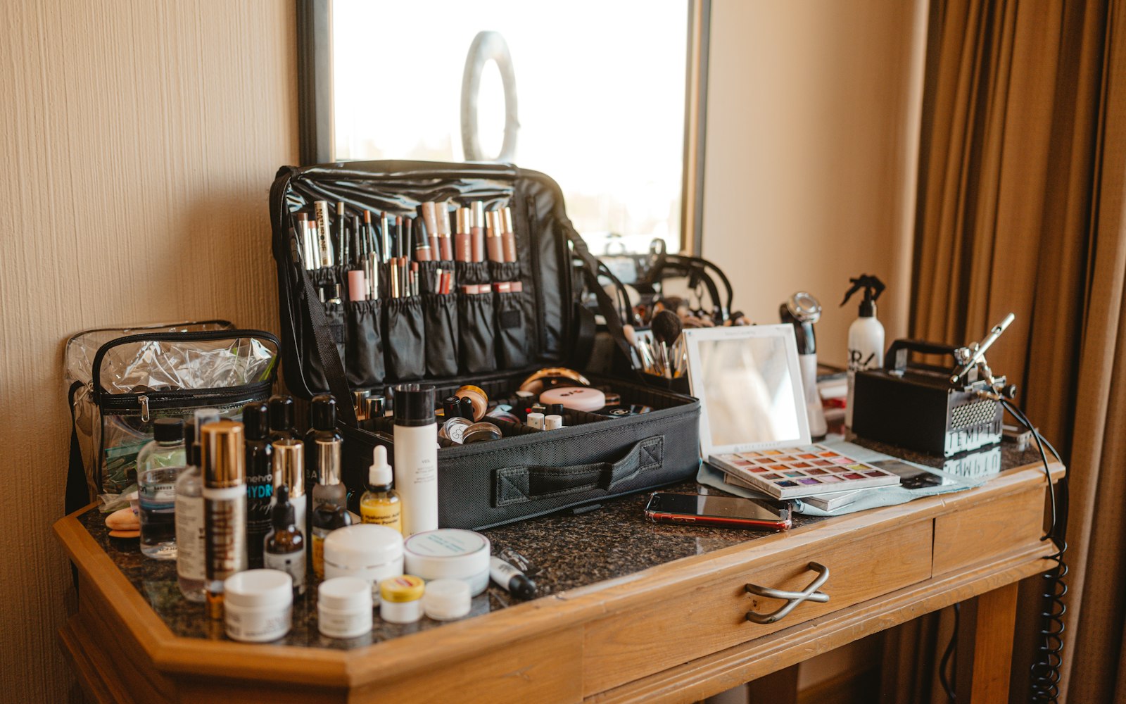 An open black makeup artist’s case of lipsticks and brushes on a wooden vanity, with skincare bottles, an eyeshadow palette, a mirror and an airbrush