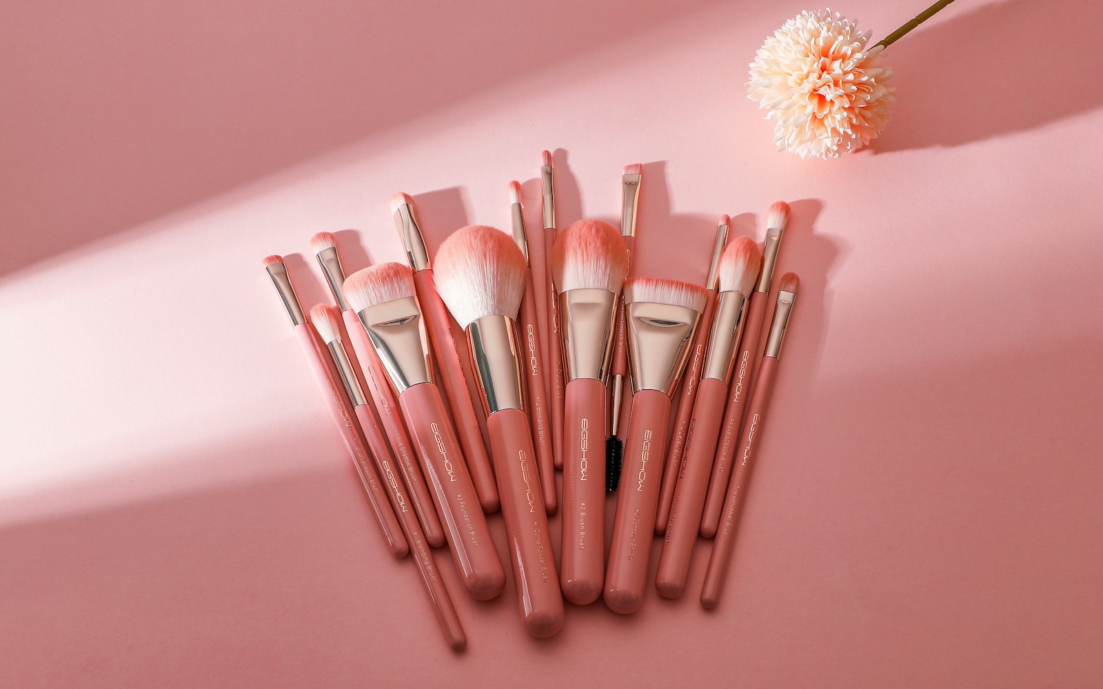 A fanned-out set of pink makeup brushes with rose-gold ferrules on a pink surface, beside a pale peach pom-pom flower in diagonal sunlight