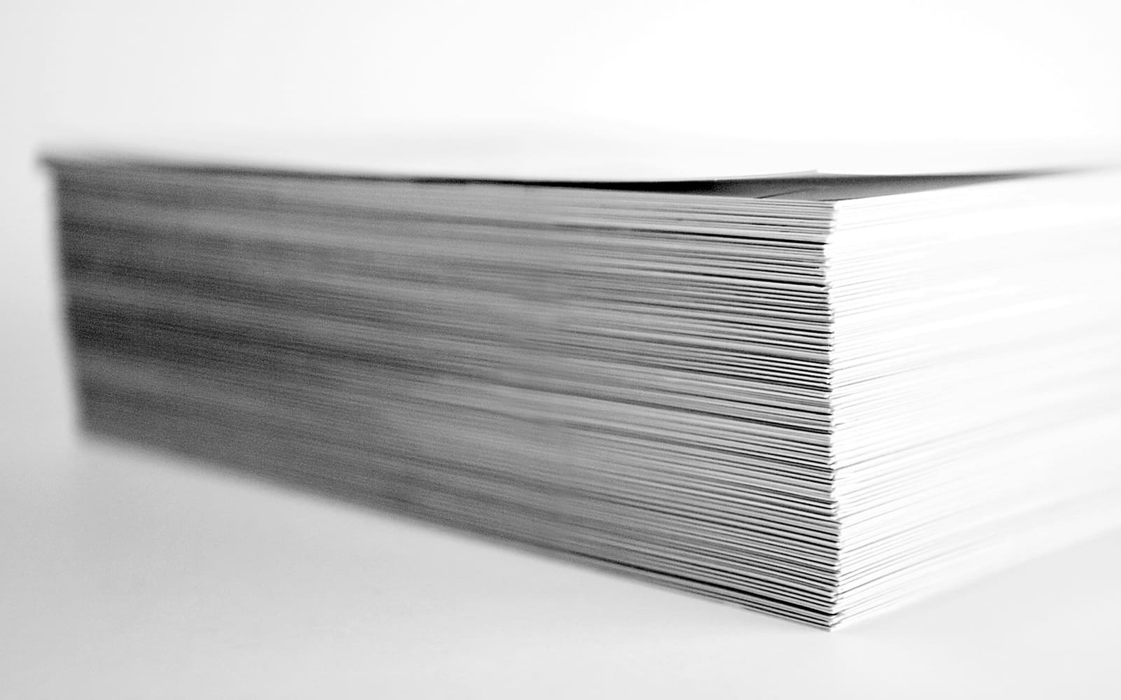 A thick stack of white paper sheets photographed side-on in black and white, the edges blurring into a plain white background