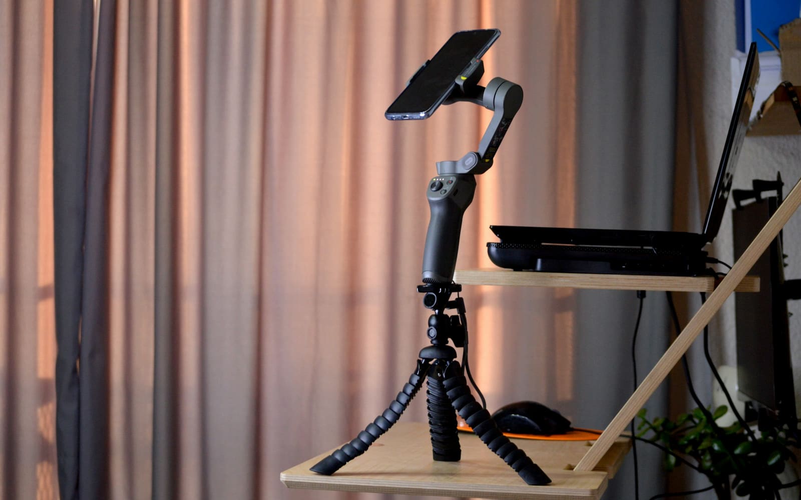 A smartphone mounted on a gray handheld gimbal stands on a small flexible tripod on a wooden desk shelf beside a laptop and pink curtains