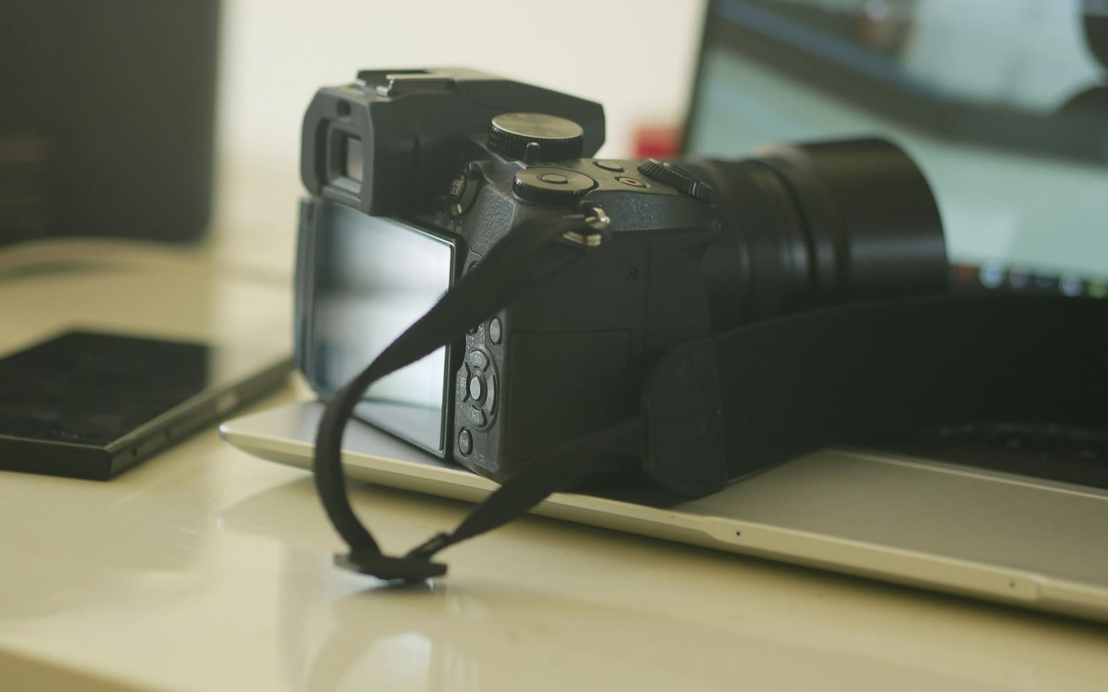A black mirrorless camera with a loose wrist strap sits on an open silver laptop beside a smartphone on a pale desk in soft daylight