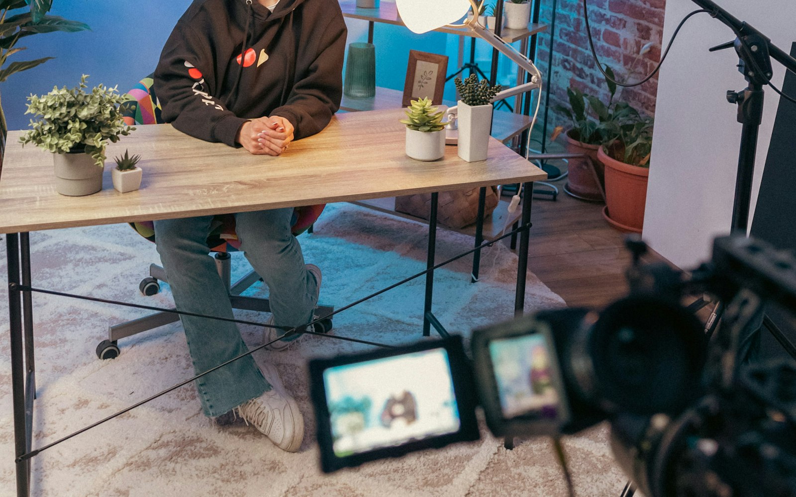 A person in a dark hoodie and jeans sits at a wooden desk with potted plants in a home studio, filmed by a video camera in the foreground