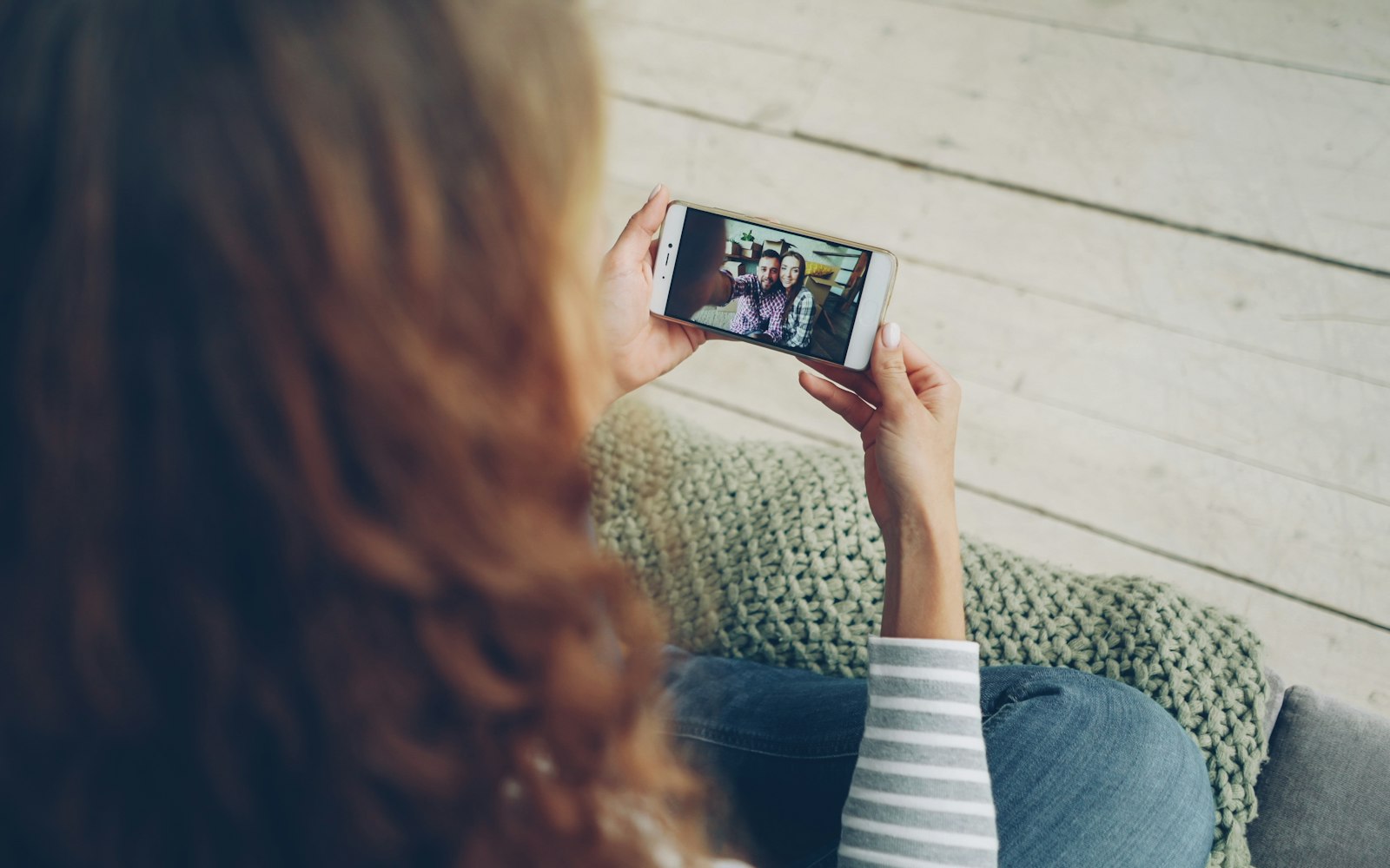 Over-the-shoulder view of a woman with long auburn hair holding a smartphone sideways, watching a video of a smiling couple, with a green knitted blanket beside her