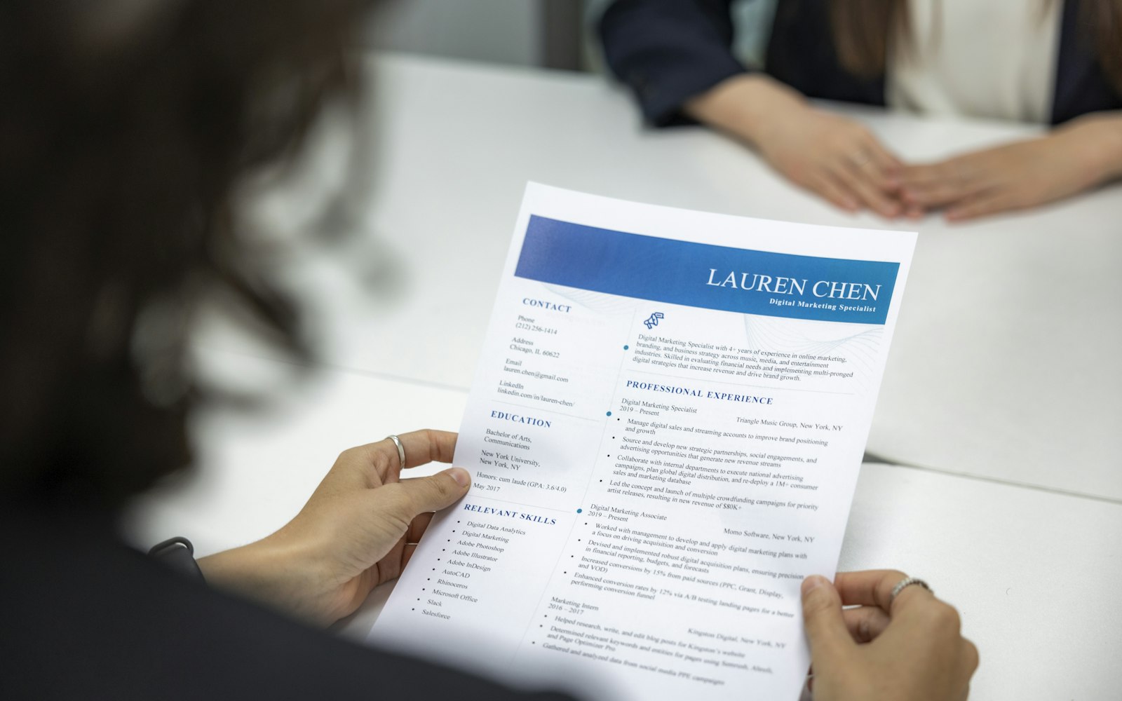 Over-the-shoulder view of a person holding a one-page résumé with a blue header and contact, education and experience sections, seated across a white table from another person