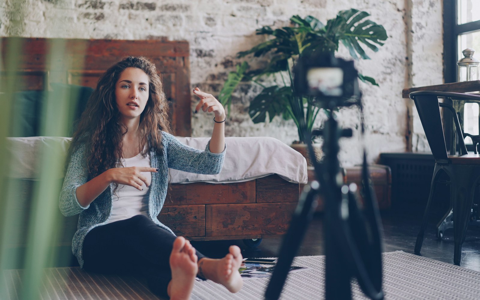 A woman with long curly hair sits on a rug by a wooden bed, talking and gesturing to a camera on a tripod in a brick-walled loft