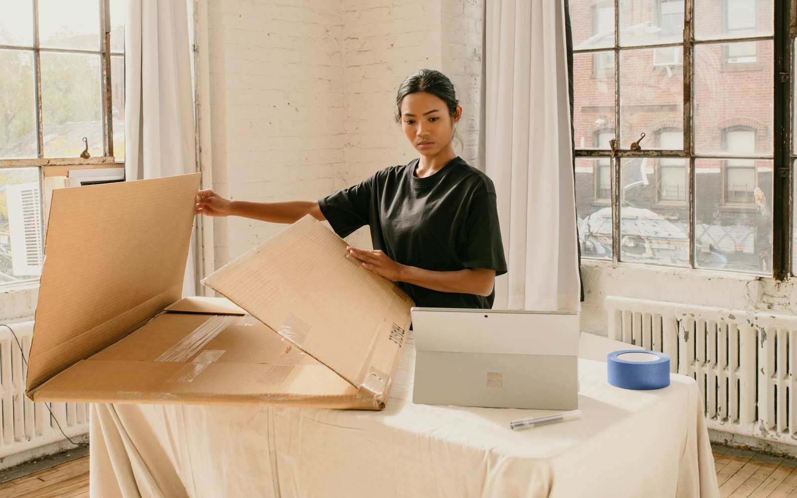 A woman in a black T-shirt opens a large flat cardboard box on a cloth-covered table beside a tablet and a roll of blue tape in a bright loft