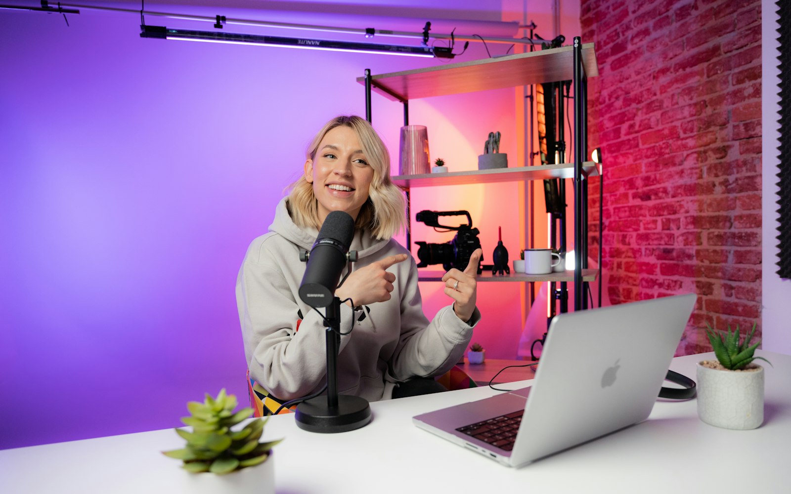 A smiling woman in a grey hoodie sits at a desk with a podcast microphone and a laptop, pointing at a camera on the shelf behind her under purple lights