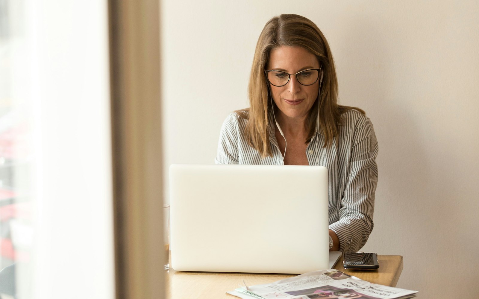 A woman in glasses and a striped shirt types on a white laptop at a wooden table, wearing earphones, with a phone and a folded newspaper beside her